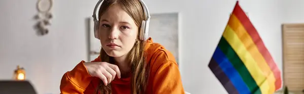 Tired young teenage girl in wireless headphones looking at camera beside pride flag, banner — Stock Photo