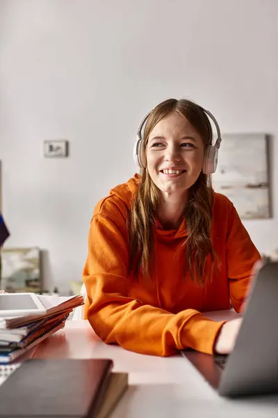 Cheerful young teenage girl in wireless headphones e-learning beside notebooks and stationery — Stock Photo