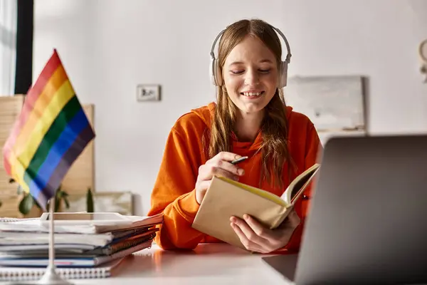 Счастливая девочка-подросток в беспроводных наушниках держит книгу рядом с ноутбуком рядом с флагом гордости и канцелярскими принадлежностями — стоковое фото