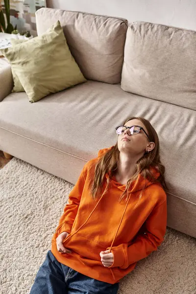 Upset teen girl in orange hoodie lounges near sofa at home, melancholy and solitude of teenager — Stock Photo