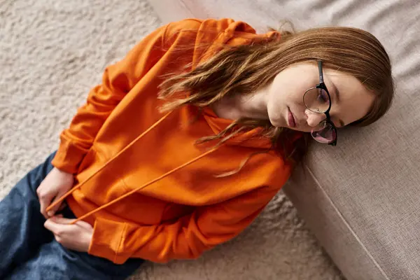 Top view of girl in orange hoodie lounges near sofa at home, melancholy and solitude of teenager — Stock Photo
