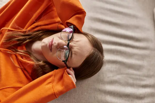 Top view of displeased teen girl in orange hoodie leaning on sofa at home, melancholy and solitude — Stock Photo