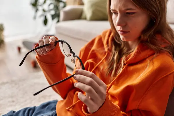 Cansada adolescente en naranja sudadera con capucha apoyada en el sofá y mirando sus gafas, melancolía y soledad - foto de stock