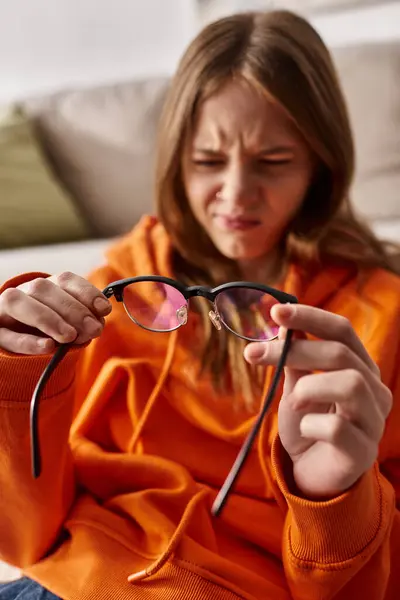 Blurred displeased teen girl in orange hoodie leaning on sofa and looking at her glasses, melancholy — Stock Photo
