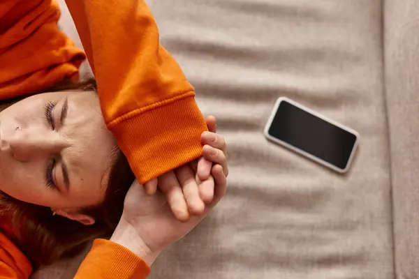 Top view of teenage girl in orange hoodie lying near smartphone on couch, stress from social media — Stock Photo