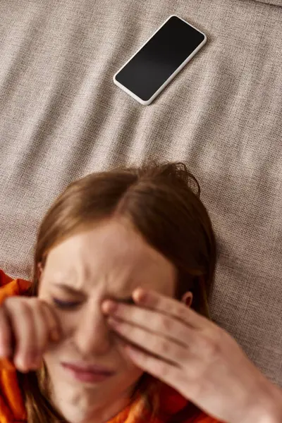 Вид зверху дівчини-підлітка в помаранчевому светрі, що лежить біля смартфона на дивані, стрес від соціальних медіа — стокове фото