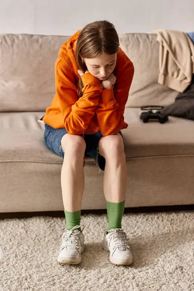 Upset teenager girl in orange hoodie sitting on couch near blurred joystick, solitude and sadness — Stock Photo
