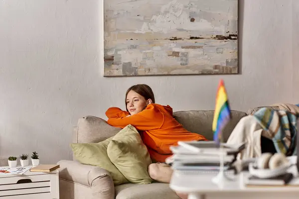 Dreamy teen girl in hoodie sits on couch with a distant look, blurred lgbtq flag on foreground — Stock Photo