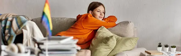 Striscione di ragazza adolescente sognante in felpa con cappuccio si siede sul divano con uno sguardo lontano, bandiera lgbtq in primo piano — Foto stock