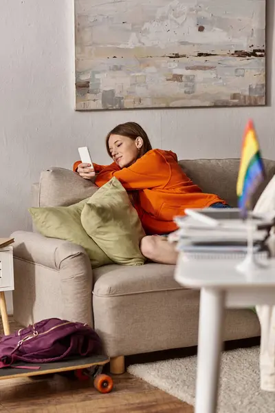 Alegre chica adolescente usando su teléfono inteligente cerca del sofá y borrosa bandera lgbt en primer plano - foto de stock