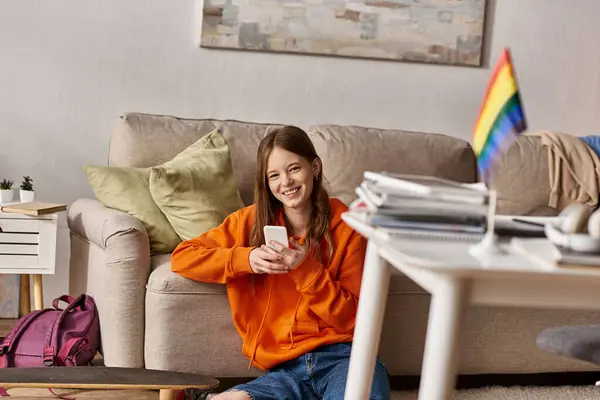 Alegre chica adolescente usando su teléfono inteligente cerca del sofá y borrosa bandera lgbt en primer plano - foto de stock