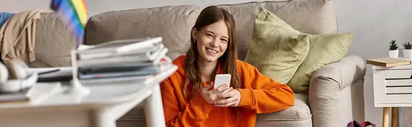 Alegre chica adolescente usando su teléfono inteligente cerca del sofá y borrosa bandera lgbt en primer plano, bandera - foto de stock