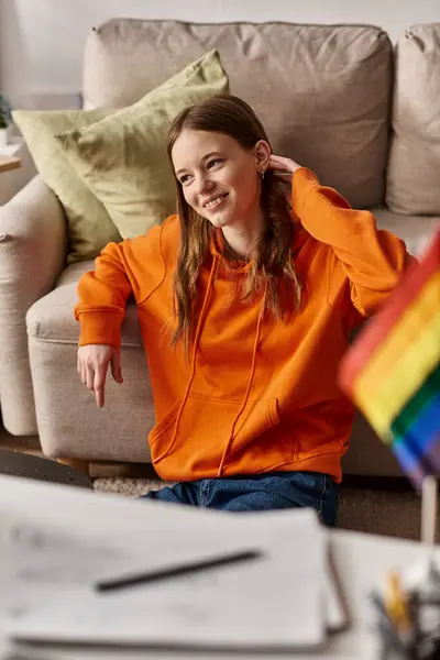 Alegre adolescente chica en naranja sudadera con capucha sentado cerca de sofá y borrosa lgbt bandera en primer plano - foto de stock