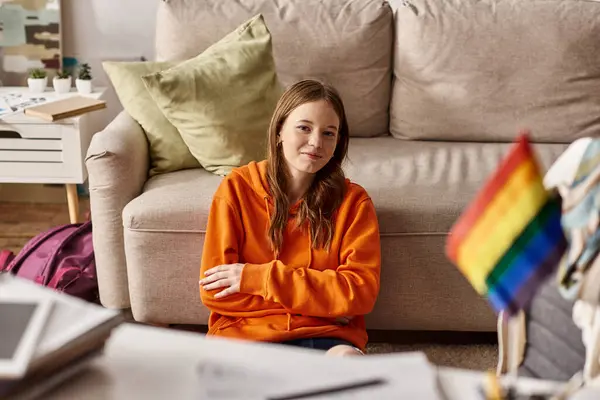 Happy teenager girl in orange hoodie sitting near couch and blurred lgbt flag on foreground — Stock Photo