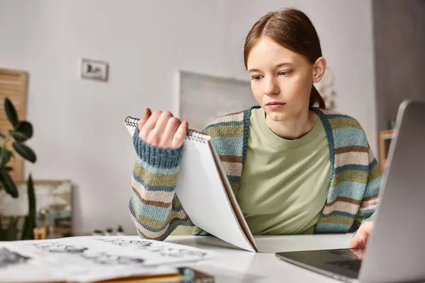 Focused teen girl looking at notebook while e-learning near laptop and sketches on desk — Stock Photo