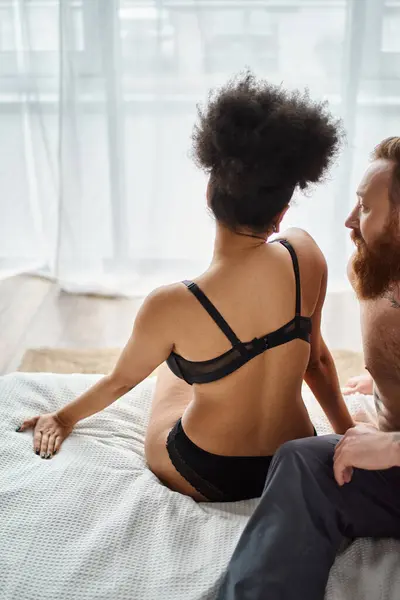 Многорасовая пара отдыхает на кровати в уютной комнате, кудрявая черная женщина в нижнем белье и бородатый мужчина — стоковое фото