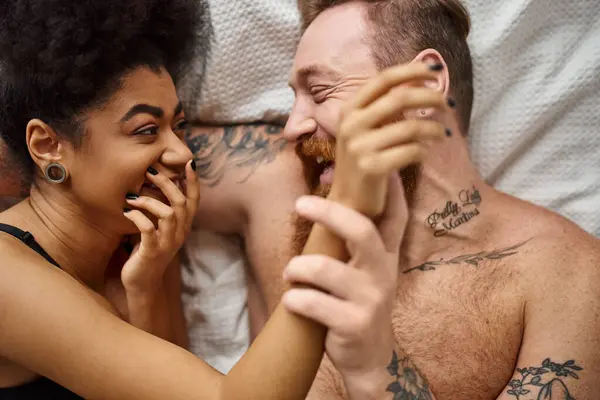 Heureuse femme afro-américaine couvrant la bouche et riant tout en étant couché sur le lit avec son petit ami, vue sur le dessus — Photo de stock