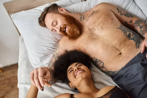 Vista dall'alto di felice uomo barbuto e tatuato sdraiato sul letto con fidanzata afro-americana, legame — Foto stock