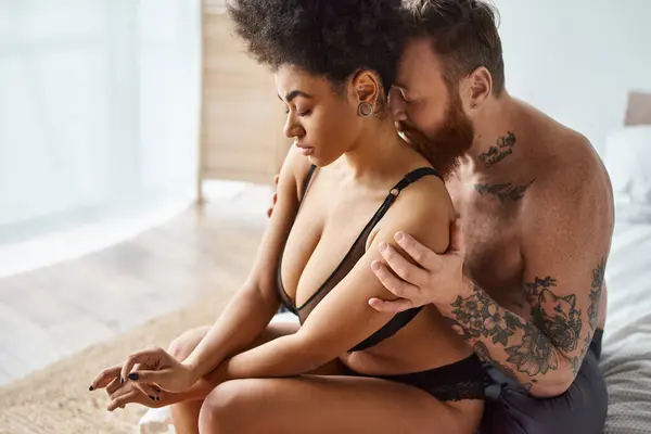 Разнообразная пара, бородатый мужчина с татуировками и черная женщина разделяют нежный момент в спальне — стоковое фото