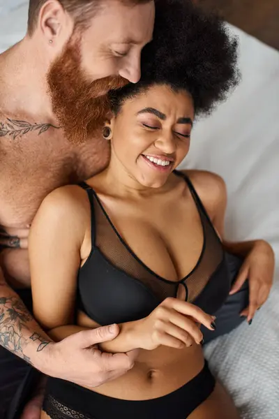 Barbudo homem com tatuagens seduzindo alegre afro-americano mulher em lingerie na cama, diverso casal — Fotografia de Stock