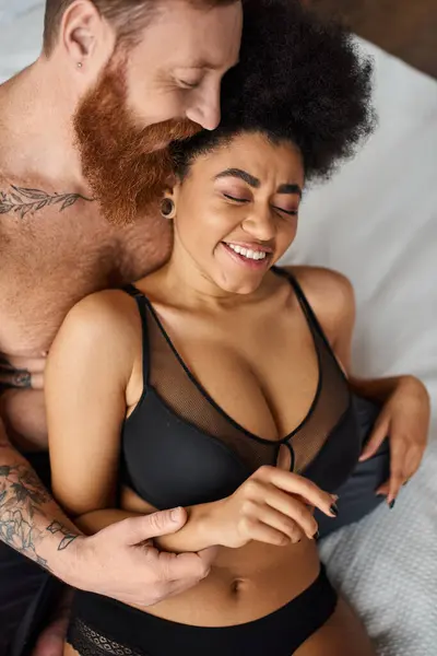 Bearded man with tattoos seducing cheerful african american woman in lingerie on bed, diverse couple — Stock Photo