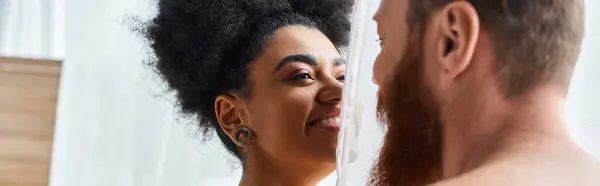 Joyful and diverse couple smiling while hiding behind curtain in a sunlit cozy bedroom, banner — Stock Photo