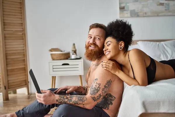 Homme barbu et femme afro-américaine, couple interracial couché sur le lit et regarder un film sur ordinateur portable — Photo de stock