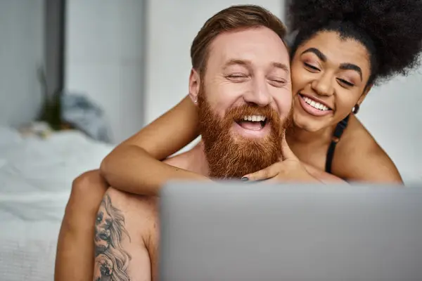 Hombre tatuado y mujer afroamericana viendo la película de comedia en el ordenador portátil y riendo juntos - foto de stock