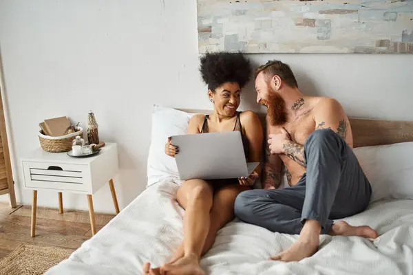 Mujer afroamericana feliz en lencería usando el ordenador portátil y acostado en la cama cerca de novio tatuado - foto de stock