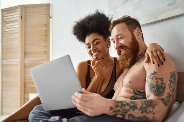 Mujer afroamericana positiva en lencería usando el ordenador portátil y acostado en la cama cerca del hombre tatuado, comedia - foto de stock
