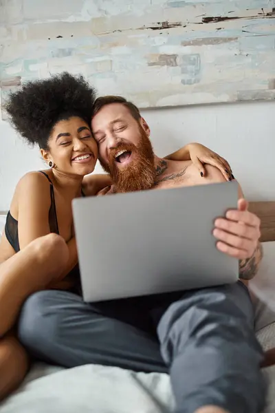 Glücklich afrikanisch-amerikanische Frau in Dessous mit Laptop und Liegen auf dem Bett in der Nähe tätowierten Mann, Film — Stockfoto