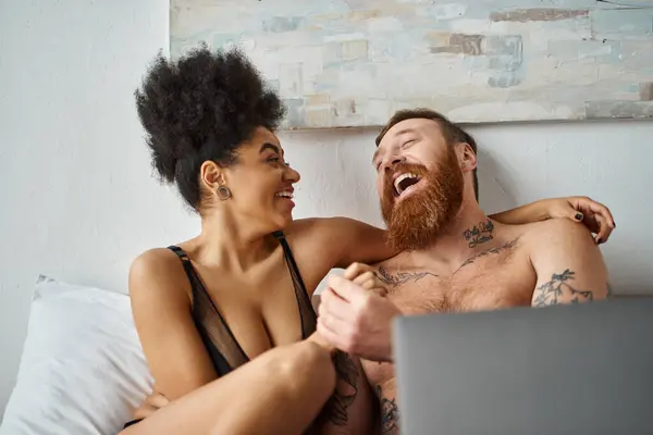 Heureuse femme afro-américaine en lingerie à l'aide d'un ordinateur portable et couché sur le lit près d'un homme tatoué, rire — Photo de stock