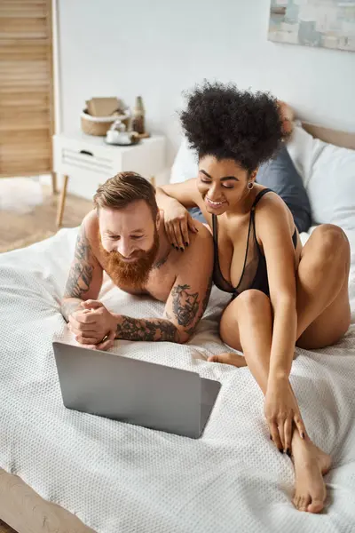 Мультикультурная пара смотрит комедийный фильм на кровати, африканская американка и бородатый татуированный мужчина — стоковое фото