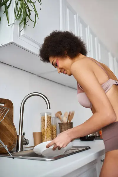 Jeune femme afro-américaine frisée en lingerie avec des taches roses sous les yeux laver la vaisselle — Photo de stock