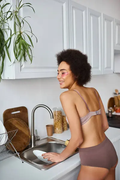 Щаслива і кучерява афроамериканка в нижній білизні з рожевими плямами під очима миття посуду — стокове фото
