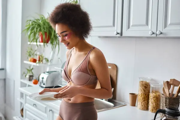 Happy and curly african american woman in lingerie with pink patches under eyes holding clean dishes — Stock Photo