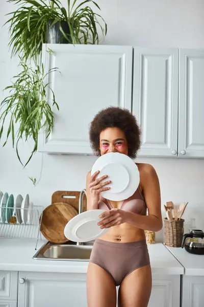 Happy african american girl in lingerie with pink patches under eyes covering face with clean dishes — Stock Photo