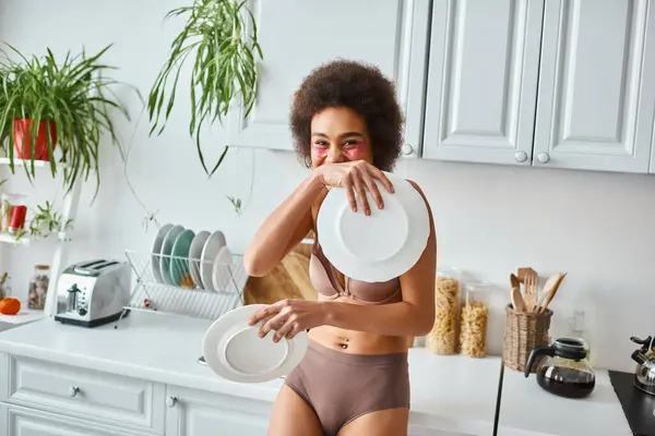 Joyeuse femme afro-américaine frisée en lingerie avec des taches roses sous les yeux tenant des plats propres — Photo de stock