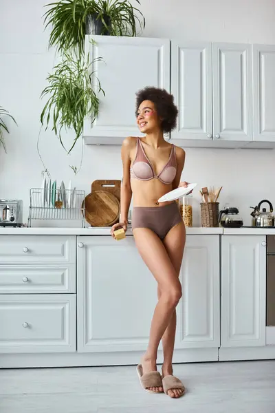 Afro-américaine en lingerie avec des taches roses sous les yeux plaque de lavage avec éponge dans la cuisine — Photo de stock