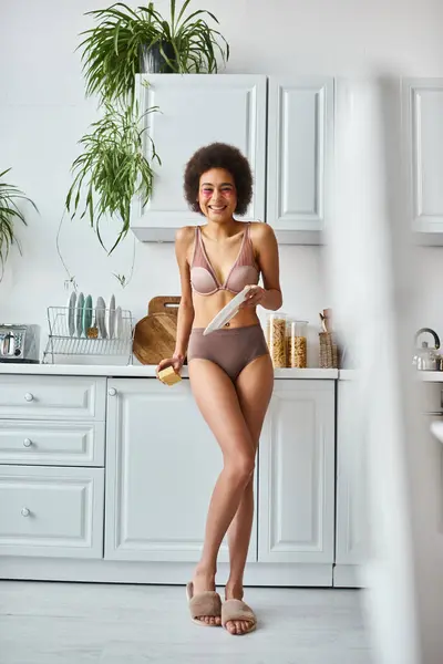 Happy black woman in lingerie with pink patches under eyes washing plate with sponge in kitchen — Stock Photo