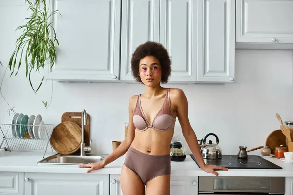 Curly african american woman in bra with pink patches under eyes standing in modern kitchen — Stock Photo