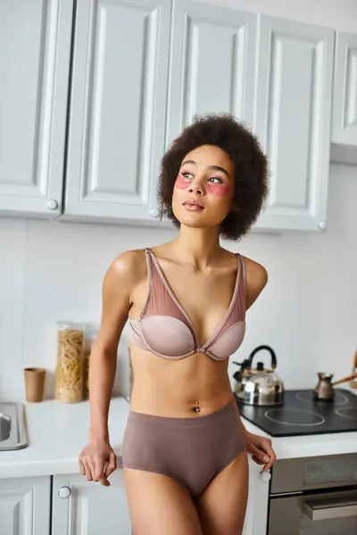Dreamy african american woman in bra with pink patches under eyes standing in modern kitchen — Stock Photo