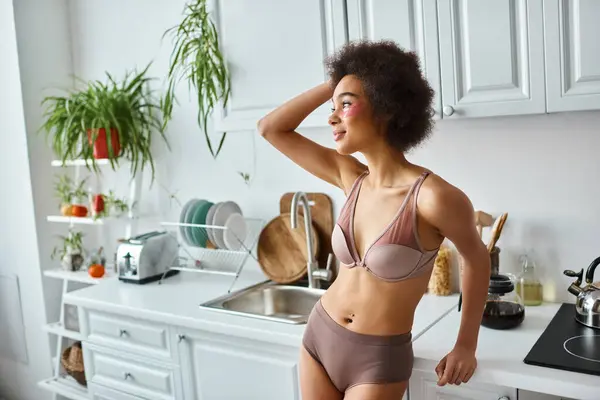 Sorridente mulher afro-americana em sutiã com manchas rosa sob os olhos em pé na cozinha moderna — Fotografia de Stock
