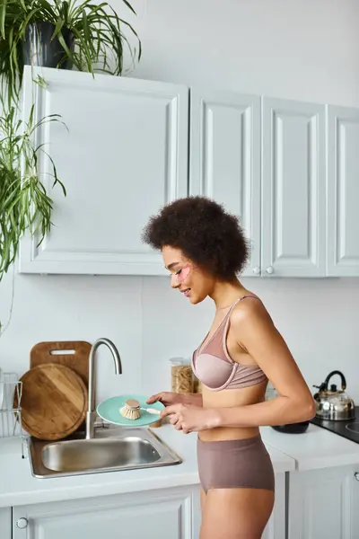 African american woman in bra with pink patches under eyes smiling and washing plate with dish brush — Stock Photo