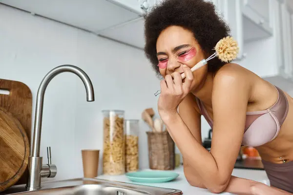 Joyful african american woman in bra with pink patches under eyes holding dish brush in kitchen — Stock Photo