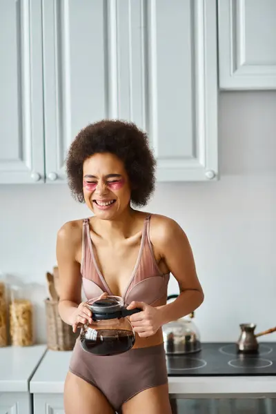 Radiante joven afroamericana mujer con parches para los ojos sosteniendo cafetera en una cocina moderna iluminada por el sol - foto de stock