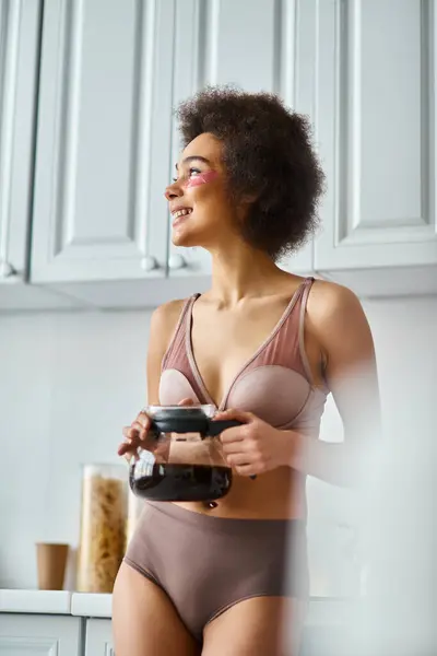 Radiant young african american woman with eye patches holding coffee pot in a sunlit modern kitchen — Stock Photo