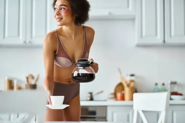 Feliz joven afroamericana mujer con parches para los ojos sosteniendo cafetera en una cocina moderna soleada - foto de stock