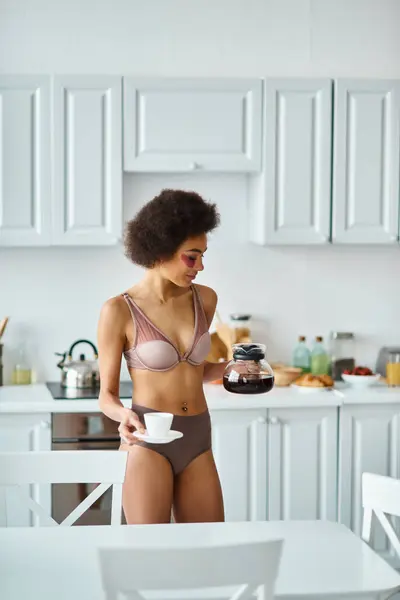 Radieuse jeune femme afro-américaine avec des patchs tenant une cafetière et une tasse dans la cuisine — Photo de stock