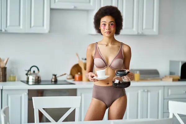 Radieuse jeune femme afro-américaine avec des patchs tenant tasse et cafetière dans la cuisine — Photo de stock